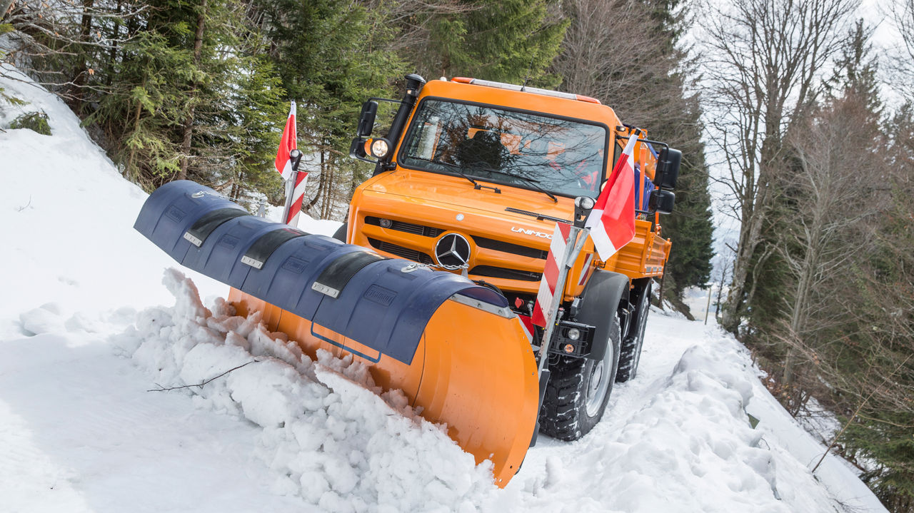 An orange Unimog with a snowplough attached clears a path in a snowy landscape. Red and white warning flags are attached to the sides of the Unimog. Snow-covered trees and a wooded slope can be seen in the background. The sky is grey and the Unimog is not covered in snow. 